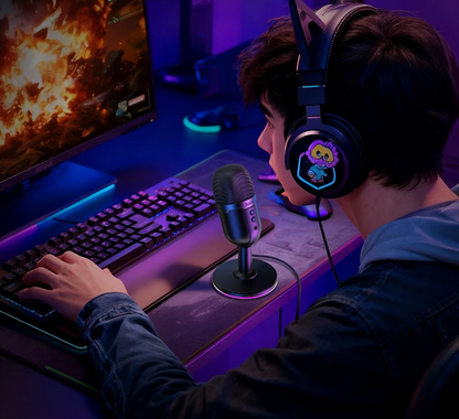A person wearing over-ear gaming headphones sits at a desk using a computer, with a microphone on a stand in front of them. The scene is lit with purple and blue ambient lighting, and the headphones feature a colorful cartoon graphic on the ear cup.