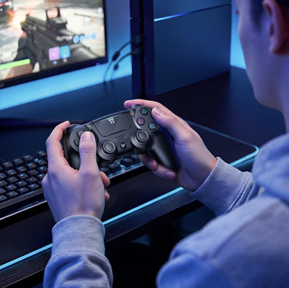 A person holding a black PlayStation 5 controller in a gaming setup, with a PS5 console standing on a desk and a monitor in the background.