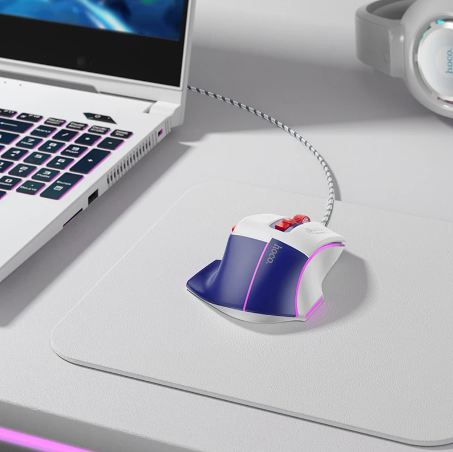 A white and blue gaming mouse with red and black accents sits on a large white desk mat, with a laptop and headphones partially visible in a clean, modern workspace setting.