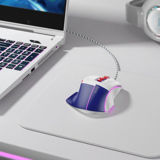 A white and blue gaming mouse with red and black accents sits on a large white desk mat, with a laptop and headphones partially visible in a clean, modern workspace setting.