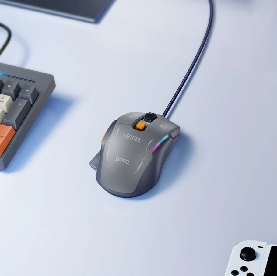 A gray HOCO S7M32 wired gaming mouse with RGB lighting and an orange scroll wheel, shown on a light blue desk next to a keyboard and controller.