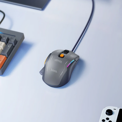 A gray HOCO S7M32 wired gaming mouse with RGB lighting and an orange scroll wheel, shown on a light blue desk next to a keyboard and controller.