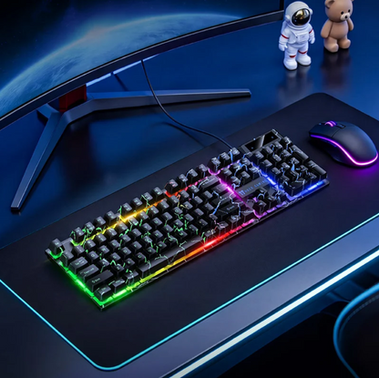 A black gaming keyboard and matching mouse set with RGB rainbow backlighting, placed on a large dark desk mat on a desk. A curved monitor stand, an astronaut figurine, and a teddy bear are visible in the scene.