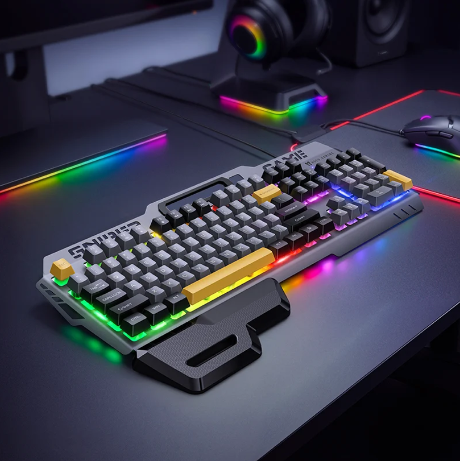 A gaming keyboard with a gray and black frame, black keycaps, and a prominent yellow accent key, shown on a dark desk with RGB lighting and a mouse in the background.