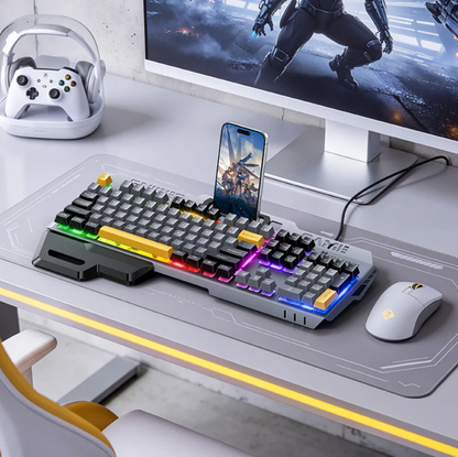 A modern gaming desk setup featuring a mechanical keyboard with RGB backlighting and colorful accent keys, a matching mouse on a large desk mat, a smartphone docked on top, a white game controller in a clear stand, and a computer monitor in a bright, clean workspace.