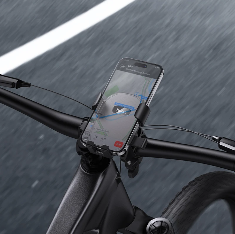 A black smartphone mounted in a clip-style bike phone holder attached to a bicycle handlebar, with a blurred road background.
